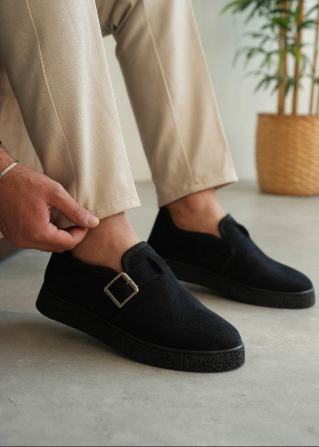 Person wearing black slip-on shoes with a buckle, sitting on a light-colored floor.